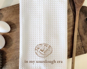 May include: White waffle weave kitchen towel with a brown illustration of a sourdough loaf of bread and the text "in my sourdough era".