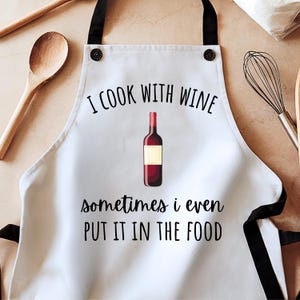 Puede incluir: Delantal blanco con correas negras y el texto "I COOK WITH WINE sometimes i even PUT IT IN THE FOOD" en letras negras. Un gráfico de botella de vino está centrado encima del texto. Una cuchara de madera y un batidor están en el fondo.
