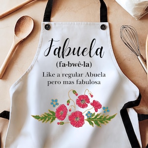 May include: White kitchen apron with black straps and a floral design. The apron has the text "Fabuela (fa-bw&ecirc;-la) Like a regular Abuela pero mas fabulosa" printed on it.