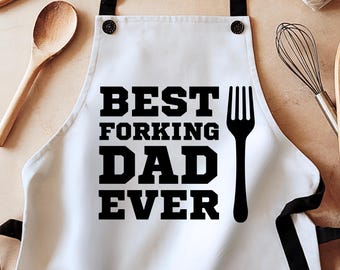 May include: White apron with black text that reads "BEST FORKING DAD EVER" and a black fork graphic. The apron has black straps and is likely used for cooking or grilling. A wooden spoon and whisk are visible in the background.
