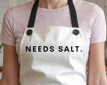 May include: White apron with black straps and black buttons. The apron has the text "NEEDS SALT." printed on the front.