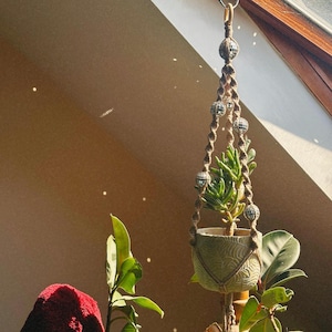 May include: A hanging macrame plant holder with a small succulent in a decorative pot. The macrame is brown with small, mirrored disco ball accents. The pot is light green with a textured design. The plant is positioned near a window.