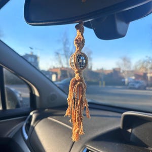May include: A tan macrame tassel with a silver disco ball hanging from the rear view mirror of a car.