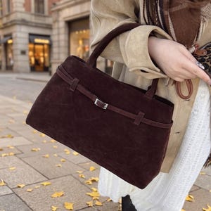 May include: A dark brown suede handbag featuring a horizontal strap across the front with a silver-toned metal buckle. The bag has two top handles and a structured rectangular shape with a soft, matte texture throughout the exterior surface.