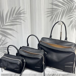 May include: Three black rectangular handbags of varying sizes, each featuring a smooth finish, a single top handle, and a silver-toned zipper closure. The bags are arranged in a row, showcasing a consistent design with a tan interior lining.