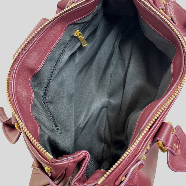 May include: A deep burgundy handbag with a smooth exterior, featuring a gold-toned metal zipper closure and matching gold hardware accents. The interior is lined with black fabric and includes a single zippered pocket with a gold-toned pull tab.