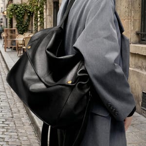 May include: A large black leather shoulder bag with a soft, slouchy design. The bag has a flap closure secured by two gold-coloured metal studs. The bag is carried over the shoulder with a long strap. The person is wearing a grey blazer.