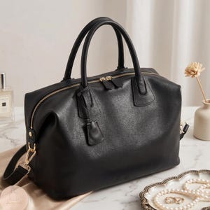 May include: A black leather handbag featuring two top handles, a gold-toned zipper closure, and a detachable shoulder strap with gold-colored metal hardware. The bag has a structured base and a smooth, matte finish with a small hanging leather tag.