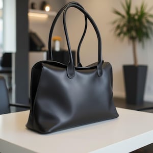 Puede incluir: Un bolso tote negro liso con un acabado mate y suave. El bolso presenta una base rectangular, una parte superior abierta y dos correas de hombro largas y delgadas unidas a los paneles frontal y posterior con costuras redondeadas.