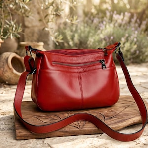 May include: A vibrant red leather crossbody bag featuring a smooth texture, a top zipper closure, and an additional horizontal zippered pocket on the front. The bag includes a matching adjustable shoulder strap attached with metal rings on both sides.