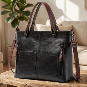 May include: Black leather tote bag with brown handles and a detachable shoulder strap. The bag features two front pockets with zippers and silver-tone hardware. The bag is sitting on a wooden surface.
