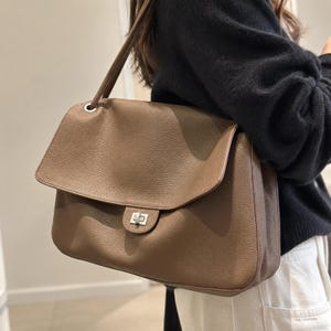 Può includere: Una borsa a spalla color tortora con texture granulata, dotata di una grande patta, una chiusura a girello in metallo argentato e una tracolla abbinata fissata con anelli in metallo argentato sugli angoli superiori della borsa.