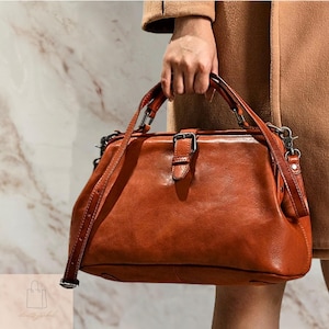 May include: A brown leather satchel bag with a top handle and a detachable shoulder strap. The bag features a buckle closure and metal hardware. The bag is a warm, reddish-brown color.