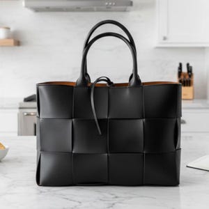 May include: A black tote bag featuring a large woven grid pattern across the exterior. The bag has two rounded top handles and a thin black tie closure at the center. The interior lining is a contrasting tan color, visible at the top edge.