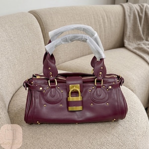 May include: A deep burgundy handbag featuring dual top handles wrapped in protective white paper, gold-tone metal hardware, and a prominent central gold padlock closure. The bag is accented with small gold studs and decorative stitching.