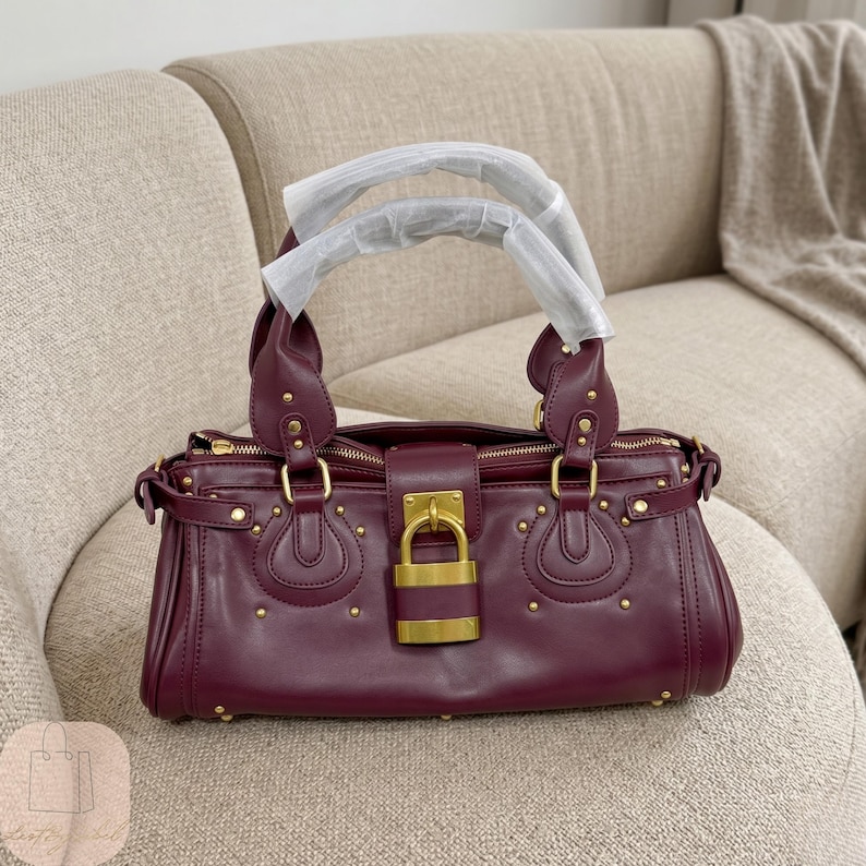 May include: A deep burgundy handbag featuring dual top handles wrapped in protective white paper, gold-tone metal hardware, and a prominent central gold padlock closure. The bag is accented with small gold studs and decorative stitching.