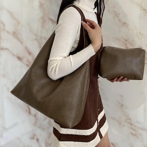 May include: A large, olive-brown tote bag with a matching small pouch. The tote has a shoulder strap and a simple, minimalist design. The pouch is rectangular with a zipper closure. The background is a marble wall.