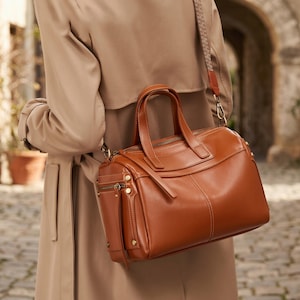 May include: A structured cognac-colored leather handbag featuring dual top handles, a zippered main compartment, and a side pocket with metallic hardware. The bag includes a detachable, patterned fabric shoulder strap with gold-toned clips.