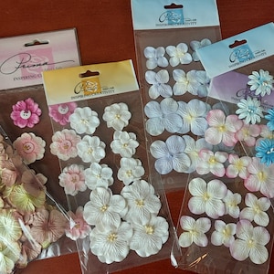 May include: Several packages of decorative paper flowers in various colors, including pink, white, blue, and lavender. The flowers are in different sizes and styles, perfect for scrapbooking or craft projects. The packages are labeled with the brand name "Prima".