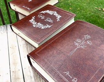 Könnte beinhalten: Vier braune, ledergebundene Journale mit weißen Nähten und dekorativen Designs. Die Journale sind in einer Reihe auf einer Holzoberfläche angeordnet. Die Designs beinhalten einen floralen Kranz mit dem Text "My Bible", einen floralen Kranz mit dem Text "Journal" und einen einzelnen floralen Stiel mit dem Text "About Day".