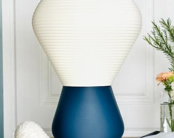 May include: A two-toned decorative vase. The base is a deep blue, and the top is a cream color with horizontal ribbed detailing. The vase is on a wooden table with a white seashell figurine.