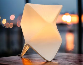 May include: A white, geometric table lamp with a warm, glowing interior. The lamp has a unique, angular design and is placed on a dark wooden surface. The background features a blurred view of a body of water and a sunset.
