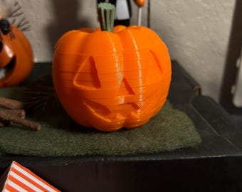 Puede incluir: Una calabaza naranja impresa en 3D con una cara tallada y un tallo verde. En el fondo, una figura temática de Halloween presenta un personaje con cabeza de calabaza en esmoquin sosteniendo un caramelo de maíz.
