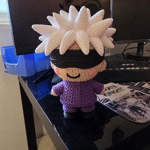 May include: A handmade, crocheted doll of a character with white spiky hair, a black blindfold, and a purple outfit. The doll has a smiling expression and is standing on a black surface. The doll is a unique, handcrafted item.