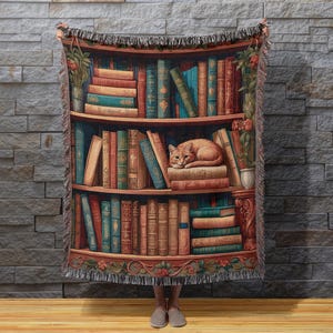 May include: A tapestry throw blanket featuring a bookshelf design with a cat resting on the books. The blanket has a fringed edge and depicts a variety of books in shades of brown, blue, and green. The background is a brick wall.