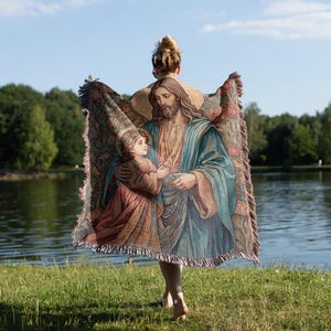 Puede incluir: Una mujer sostiene una manta de tapiz con una imagen religiosa de Jes&uacute;s y un ni&ntilde;o. El tapiz est&aacute; tejido con intrincados detalles y colores vibrantes. El fondo es una imagen borrosa de un lago y &aacute;rboles.