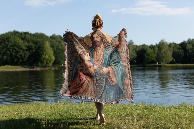Puede incluir: Una mujer sostiene una manta de tapiz con una imagen religiosa de Jes&uacute;s y un ni&ntilde;o. El tapiz est&aacute; tejido con intrincados detalles y colores vibrantes. El fondo es una imagen borrosa de un lago y &aacute;rboles.
