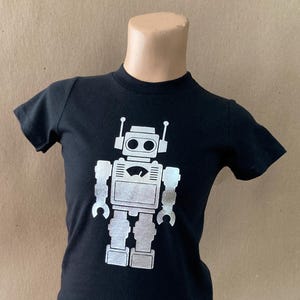 May include: Black t-shirt with a silver robot graphic. The robot has a round head with two eyes and antennas.