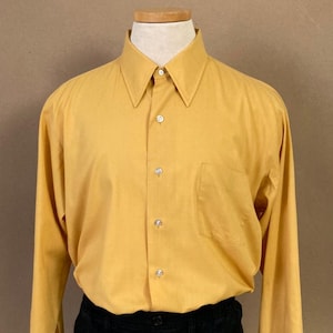 May include: Mustard yellow button-down shirt with a pointed collar and a chest pocket. The long-sleeved shirt is paired with black shorts. The shirt has a classic design.