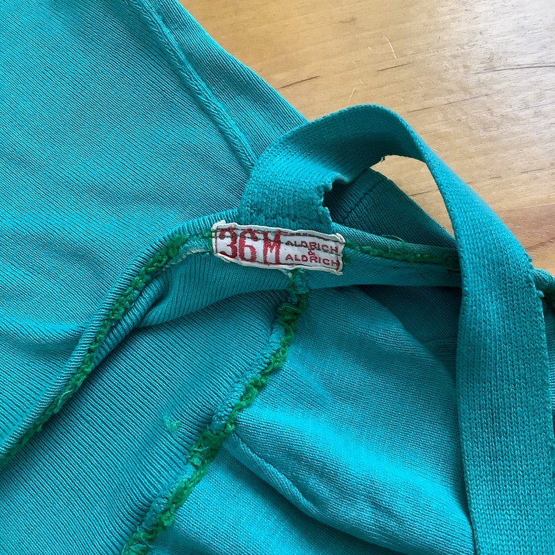 Pu&ograve; includere: Primo piano di una camicia blu turchese con una cucitura verde e un'etichetta bianca che recita "36M Aldrich".