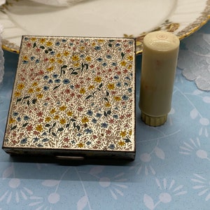 May include: A vintage floral patterned compact with a matching lipstick tube. The compact is made of metal and has a hinged lid. The lipstick tube is made of plastic and has a gold-colored cap.