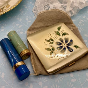 Op de afbeelding: Een vintage verguld compact met een bloemenontwerp in tinten blauw, groen en goud. Het compact bevindt zich in een bruine fluwelen zak en wordt vergezeld door twee lippenstiftbuizen, een blauwe en een groene.