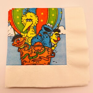 May include: A square paper napkin featuring a colorful illustration of Big Bird, Cookie Monster, Bert, and Ernie riding in a hot air balloon. The balloon is orange, green, and blue with stars. The word COOKIES is printed on the balloon basket.