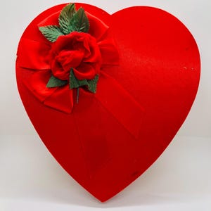May include: A red heart-shaped box with a velvet finish. It features a red satin bow and a fabric rose with green leaves. This box is suitable for gifts or special events.