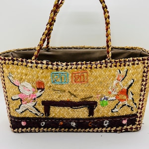 May include: Woven straw tote bag with embroidered bunny figures playing pool. The bag features a braided handle and a dark brown trim with colorful accents. The interior is lined with a brown fabric.