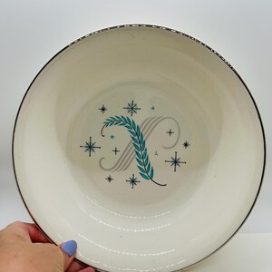 May include: Cream-colored ceramic bowl with a silver rim. The bowl features a stylized letter "X" with a teal leaf design and gray stripes, surrounded by teal and black starbursts. A hand is holding the bowl.