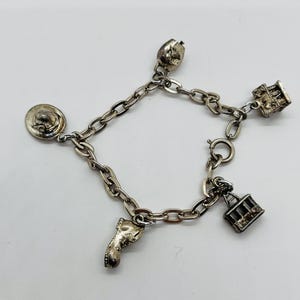 May include: A silver-toned metal charm bracelet featuring four distinct dangling charms: a round hat, a small boot, a cable car, and a rectangular building structure. The bracelet is constructed with oval-shaped links and includes a circular spring ring clasp.