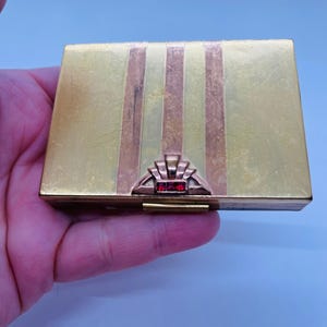May include: A rectangular, gold-toned compact with vertical stripes in gold and copper. The clasp features a decorative fan design with red accents. The compact is likely vintage and shows signs of wear.
