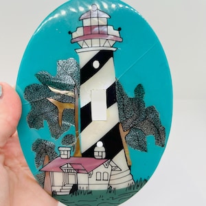 May include: Oval light switch cover featuring a lighthouse design. The lighthouse has black and white stripes, a red roof, and is set against a teal background. Trees and grass are also depicted.