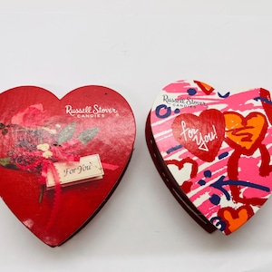 May include: Two heart-shaped Russell Stover Candies boxes. One is red with a floral design and a "For You" tag. The other is pink, red, and white with hearts and the words "for you!"