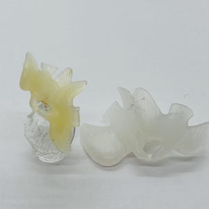 May include: Two bird-shaped decorative objects. One is a light yellow color and mounted on a clear, textured base. The other is a translucent white color. Both have a carved appearance and are set against a white background.