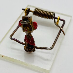 May include: A vintage-style mouse trap with a decorative mouse figurine. The mouse has a gold-colored body, red ears, and a red jacket. The trap is made of metal and sits on a clear, rectangular base.
