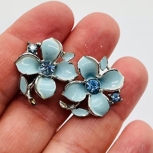 Puede incluir: Un par de pendientes en forma de flor azul claro con detalles metálicos plateados. Cada pendiente presenta una piedra preciosa azul central y piedras de acento más pequeñas. Los pendientes tienen un diseño floral con pétalos y hojas de esmalte azul claro.