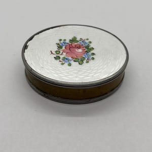 May include: An oval-shaped trinket box with a white enamel lid. The lid features a pink rose and blue floral design. The box has a silver-colored rim and a gold-colored base. A decorative item.