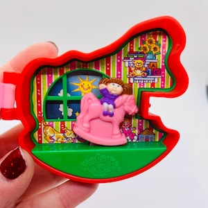 May include: A vintage Cabbage Patch Kids toy in a red rocking horse shape. The toy features a pink rocking horse with a doll, a window with a sun, and a shelf with a teddy bear and flowers. The base is green.