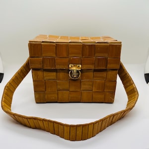 May include: A rectangular bamboo handbag with a woven pattern. The bag features a gold-colored clasp and a matching strap. The bag is made of light brown bamboo and has a boxy shape.